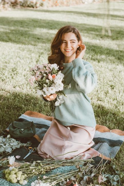 A young woman with wavy brown hair sitting on a patterned blanket on a grassy field, holding a lush floral bouquet featuring white, pink, and purple flowers with greenery. She is wearing a light blue 