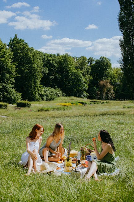 A group of three women enjoying a picnic on a lush, green meadow in Richmond Park, surrounded by tall trees and vibrant foliage. They are seated on a white blanket, with an assortment of flowers, a wi