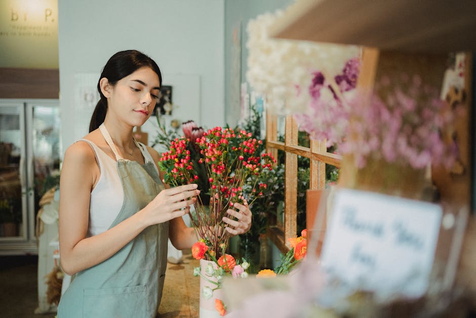 A young female florist with dark hair, wearing a beige apron over a white sleeveless top, arranging a bouquet of vibrant pink and red flowers with green stems. The flowers are displayed on a wooden co
