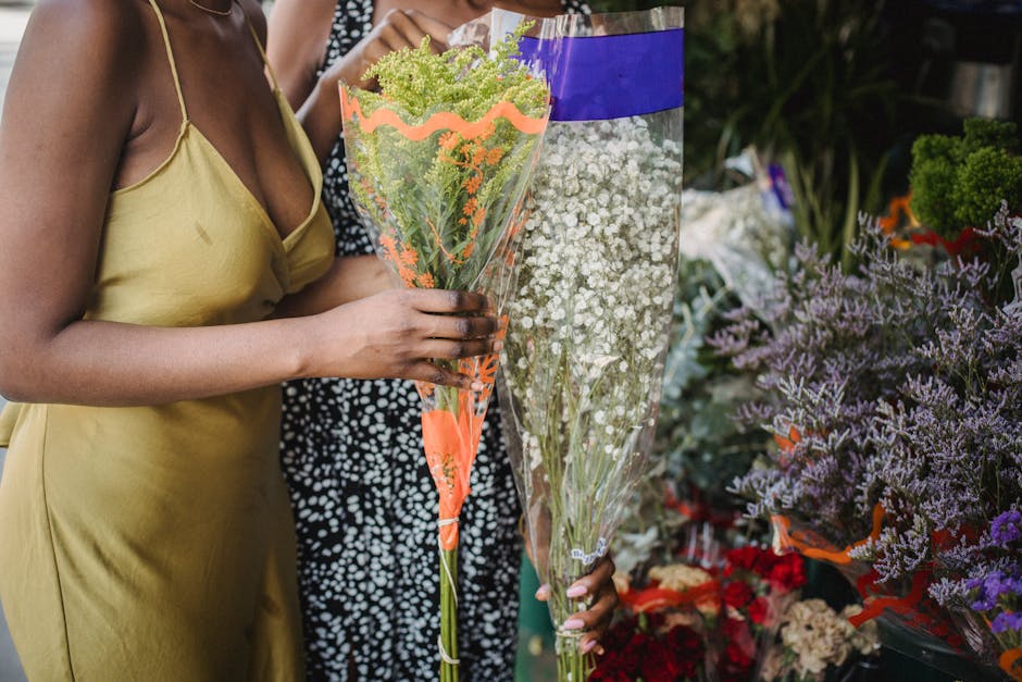 A woman wearing a yellow satin slip dress is holding a bouquet of small, delicate green flowers with rounded clusters, wrapped in clear cellophane with orange accents, at a flower shop. The bouquet is